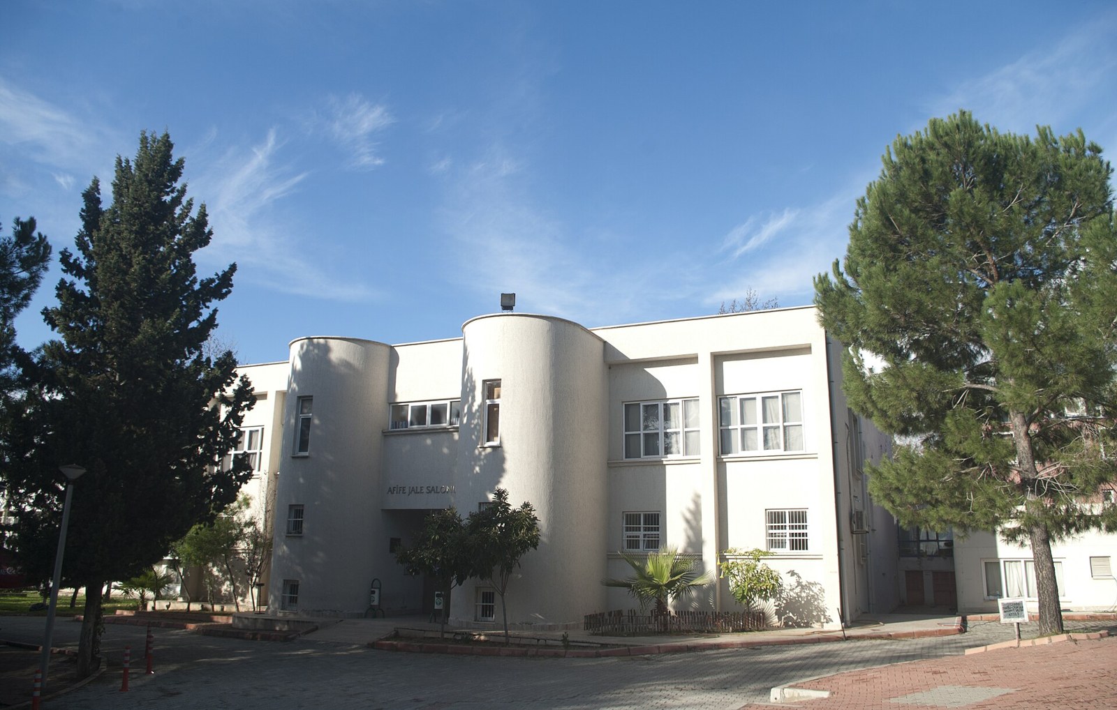 White two-storey building with rounded towers, signed Afife Jale Salonu, among pine trees under a blue sky