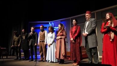 Nine actors in late Ottoman-era costumes, some wearing fezzes, stand on a stage while one speaks into a microphone.