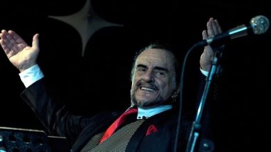 Elderly man in a dark suit and red tie smiling on stage with arms outstretched beside a microphone