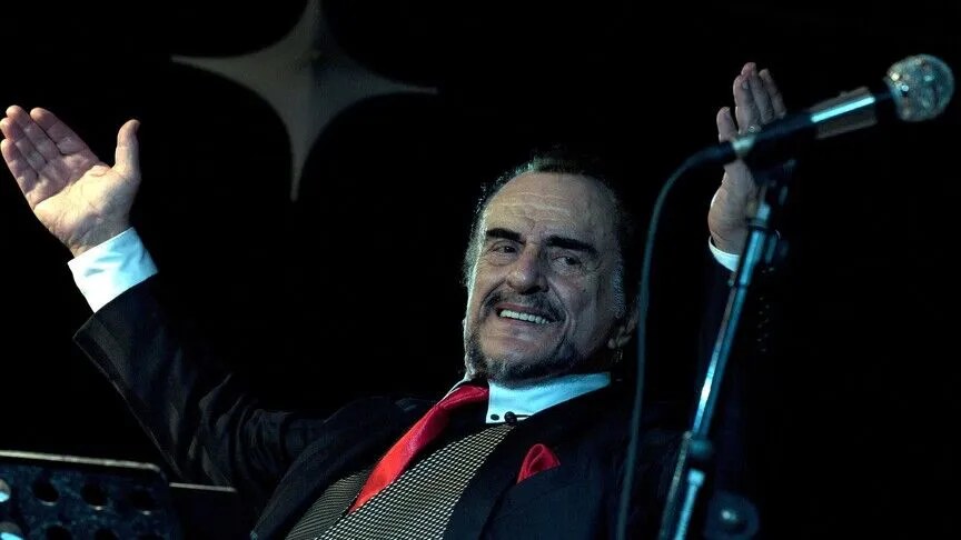 Elderly man in a dark suit and red tie smiling on stage with arms outstretched beside a microphone