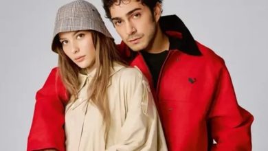 A young woman in a plaid bucket hat and beige coat poses beside a young man in a red jacket against a plain grey backdrop.