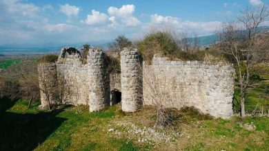 Ruined stone fortress walls and round towers on a grassy hilltop, with a valley and mountains in the distance.