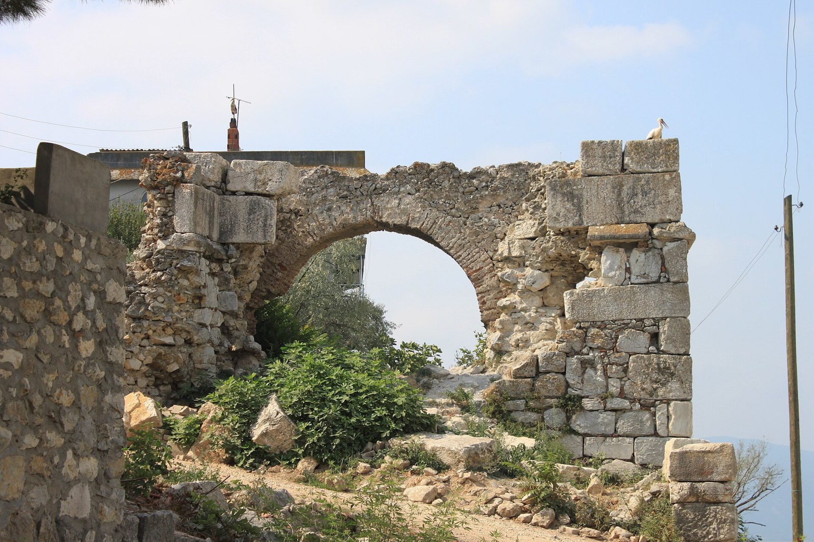Ruined ancient stone arch with a stork perched on top, surrounded by shrubs and village buildings under a hazy sky