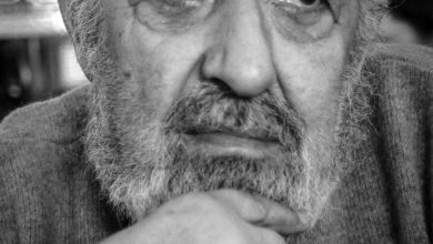 Black-and-white close-up portrait of an elderly bearded man in a wool sweater, resting his chin on his hand and looking aside.