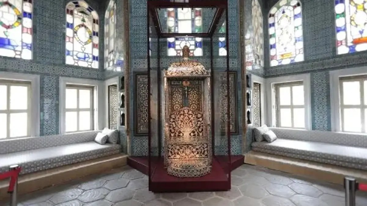 Ornate mother-of-pearl inlaid throne in a glass display case in a tiled chamber with stained-glass windows and cushioned benches