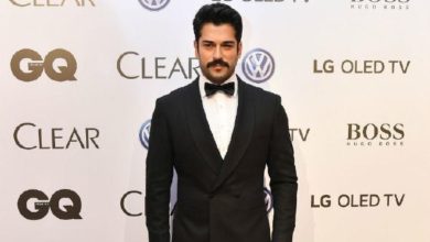 Man with dark hair and a mustache in a black tuxedo and bow tie stands in front of a sponsor backdrop at an event