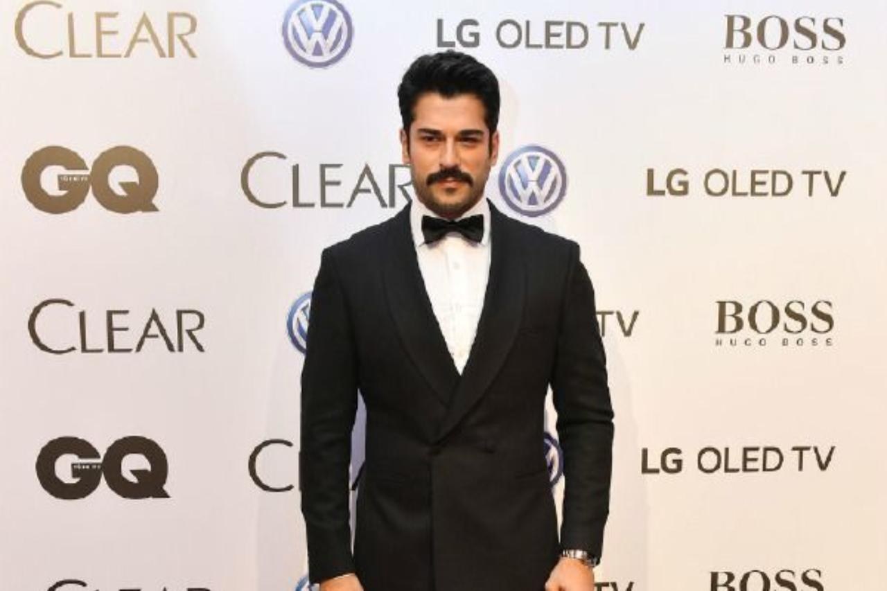 Man with dark hair and a mustache in a black tuxedo and bow tie stands in front of a sponsor backdrop at an event
