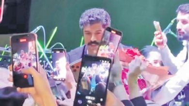 Curly-haired man with a moustache on stage, surrounded by fans holding up smartphones to film him at a concert