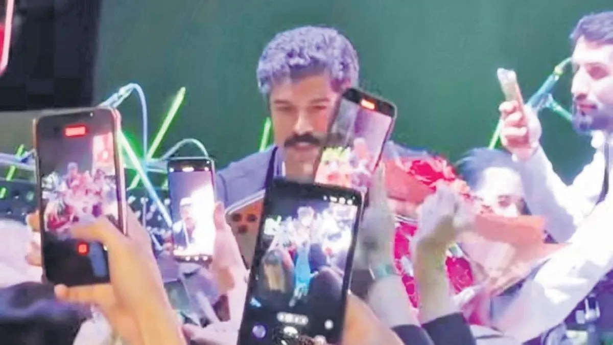 Curly-haired man with a moustache on stage, surrounded by fans holding up smartphones to film him at a concert