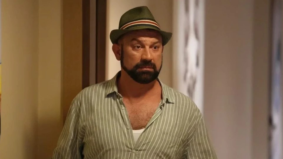 Bearded man in a green fedora and a green striped shirt stands in an indoor hallway in a still from a TV series.