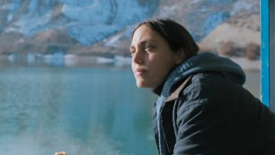A young woman in a dark hooded jacket holds a pastry and gazes out over a turquoise lake with snowy mountains behind her.