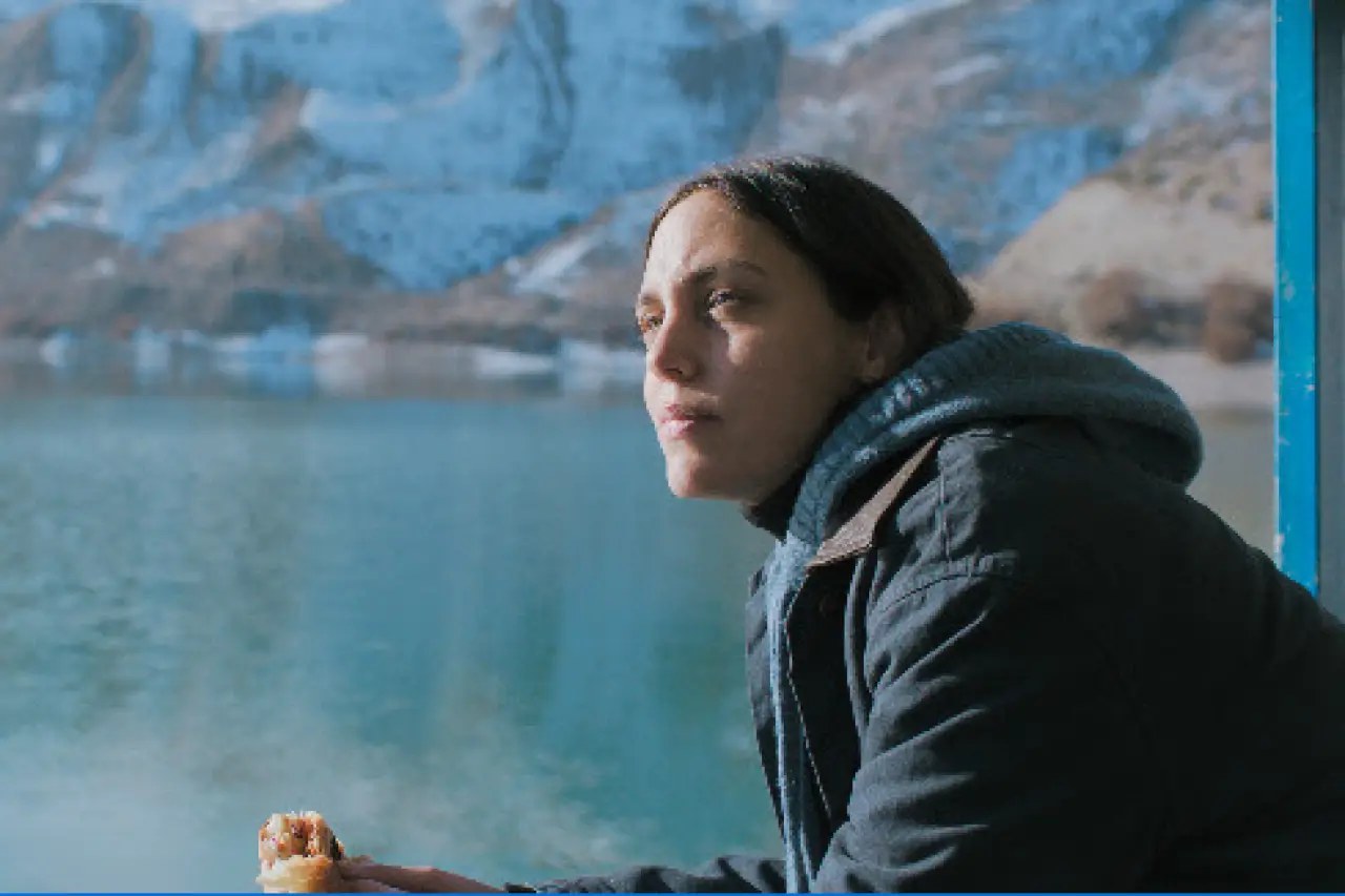 A young woman in a dark hooded jacket holds a pastry and gazes out over a turquoise lake with snowy mountains behind her.