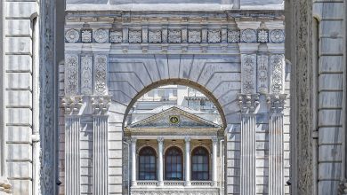 Ornate white marble gateway with columns and lamps framing an arched view of a palace facade, in the style of Dolmabahçe.