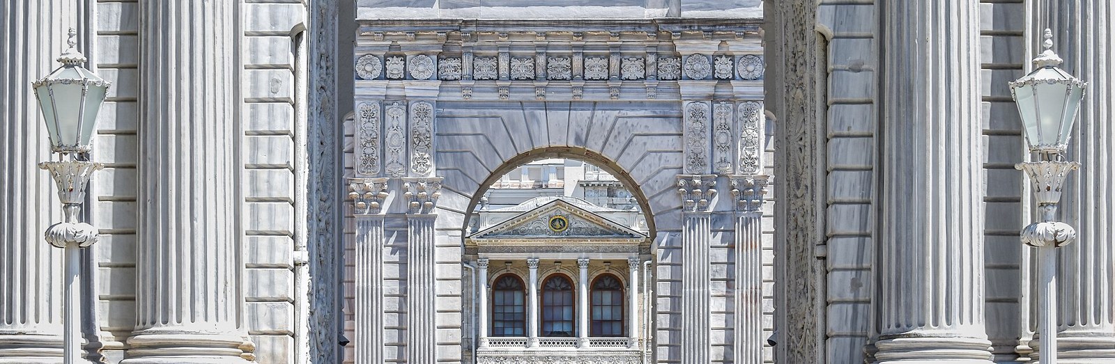 Ornate white marble gateway with columns and lamps framing an arched view of a palace facade, in the style of Dolmabahçe.