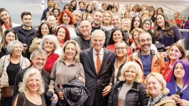 An elderly man in a dark suit and orange tie smiles in the middle of a large, cheerful crowd of mostly women posing indoors.
