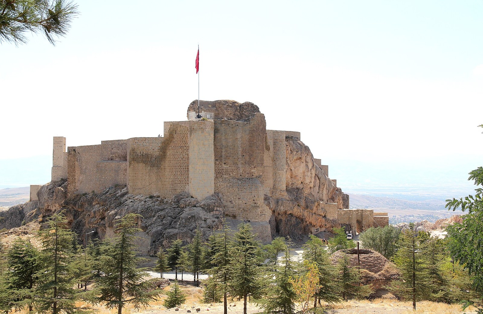 Harput Castle, a stone fortress on a rocky hilltop with a red Turkish flag, surrounded by pine trees and a wide valley behind.