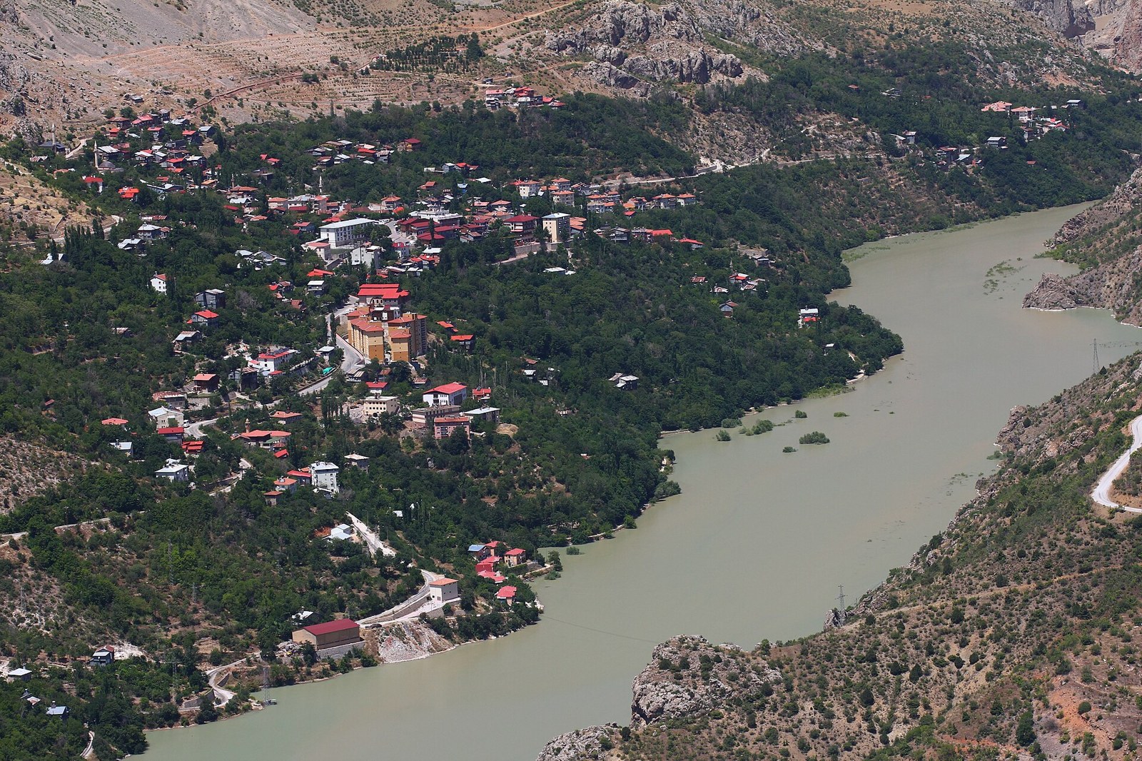 Aerial view of a hillside town with red-roofed houses beside a wide green river in a rocky mountain canyon