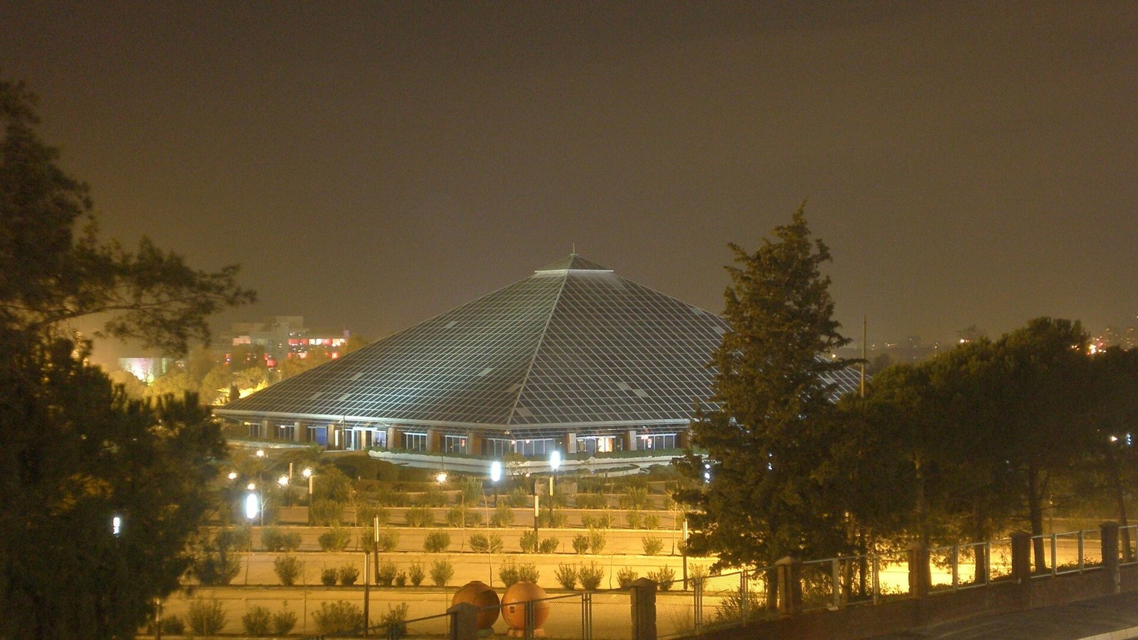 Large glass pyramid-roofed building lit up at night, seen through trees, with illuminated grounds in the foreground
