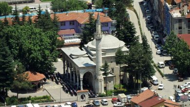 Aerial view of an Ottoman-style mosque with a single minaret and domes, with worshippers praying outside, in a Turkish town