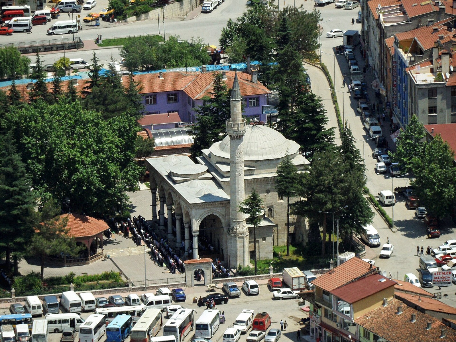 Aerial view of an Ottoman-style mosque with a single minaret and domes, with worshippers praying outside, in a Turkish town