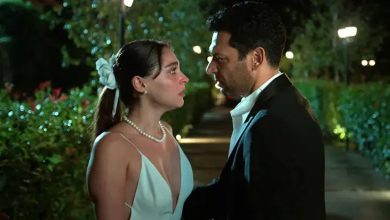 A woman in a white dress with a pearl necklace and a man in a dark suit face each other closely in a lit garden at night.