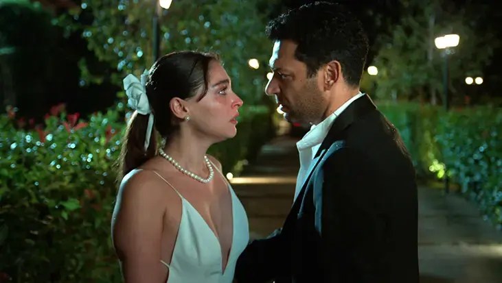 A woman in a white dress with a pearl necklace and a man in a dark suit face each other closely in a lit garden at night.