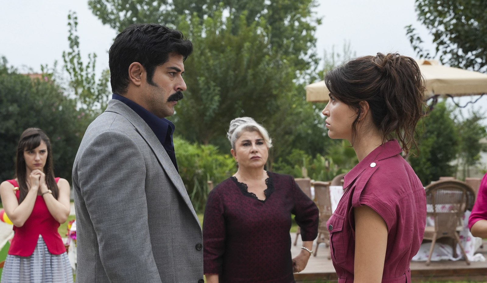 A man in a gray suit and a woman in a magenta dress face each other tensely in a garden while two women watch.