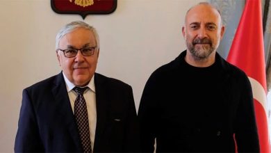 Two men pose side by side indoors: one older in a suit and glasses, one bald and bearded in black, with a Turkish flag beside them.