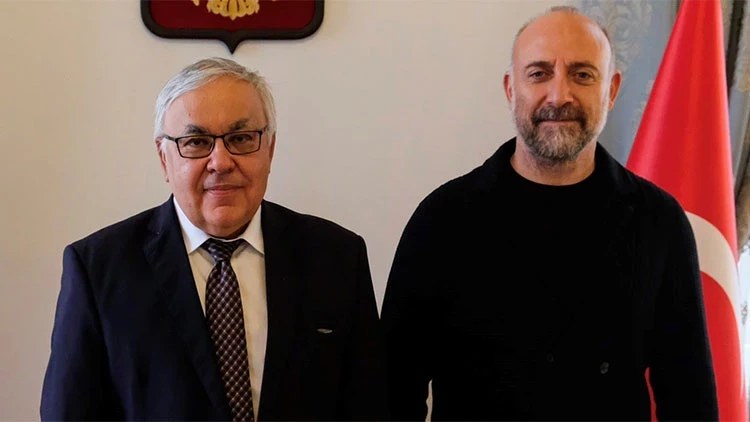Two men pose side by side indoors: one older in a suit and glasses, one bald and bearded in black, with a Turkish flag beside them.