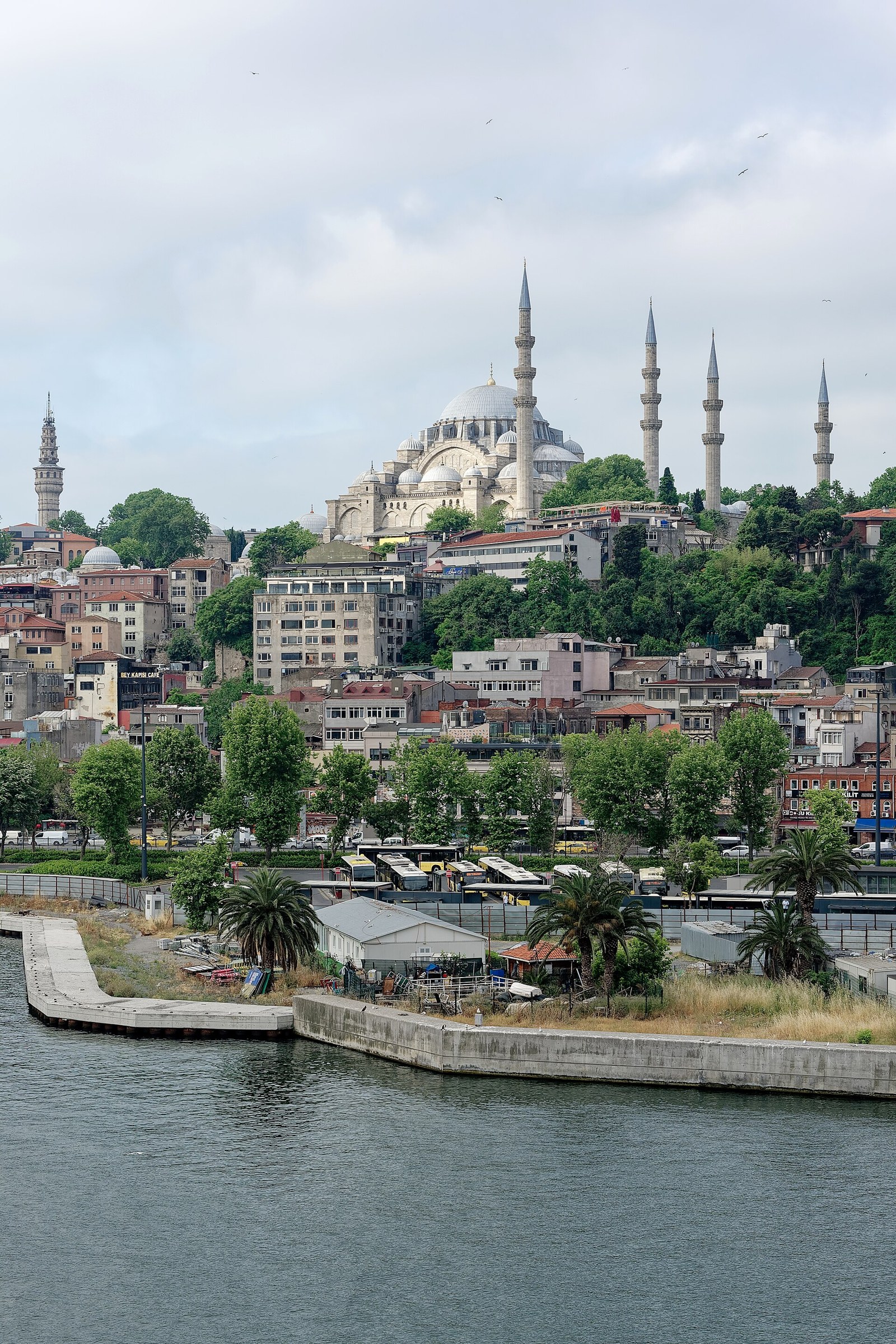 Large domed mosque with four minarets on a wooded hillside above a waterfront neighborhood, with a tower at left under a cloudy sky