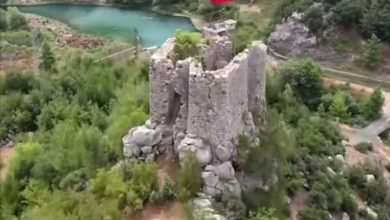 Aerial view of a ruined stone castle tower on a rocky hilltop with a Turkish flag, surrounded by forest and a turquoise lake.