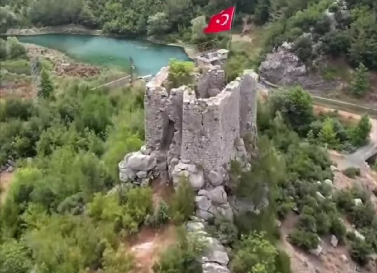 Aerial view of a ruined stone castle tower on a rocky hilltop with a Turkish flag, surrounded by forest and a turquoise lake.