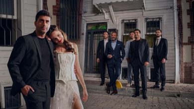 A man in a black suit stands beside a woman in a white corset dress. Five men in suits stand behind them on a cobblestone street.