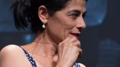 Woman with dark hair in a ponytail and a navy polka-dot top smiles, hand at her chin, on a stage with a dark blue backdrop