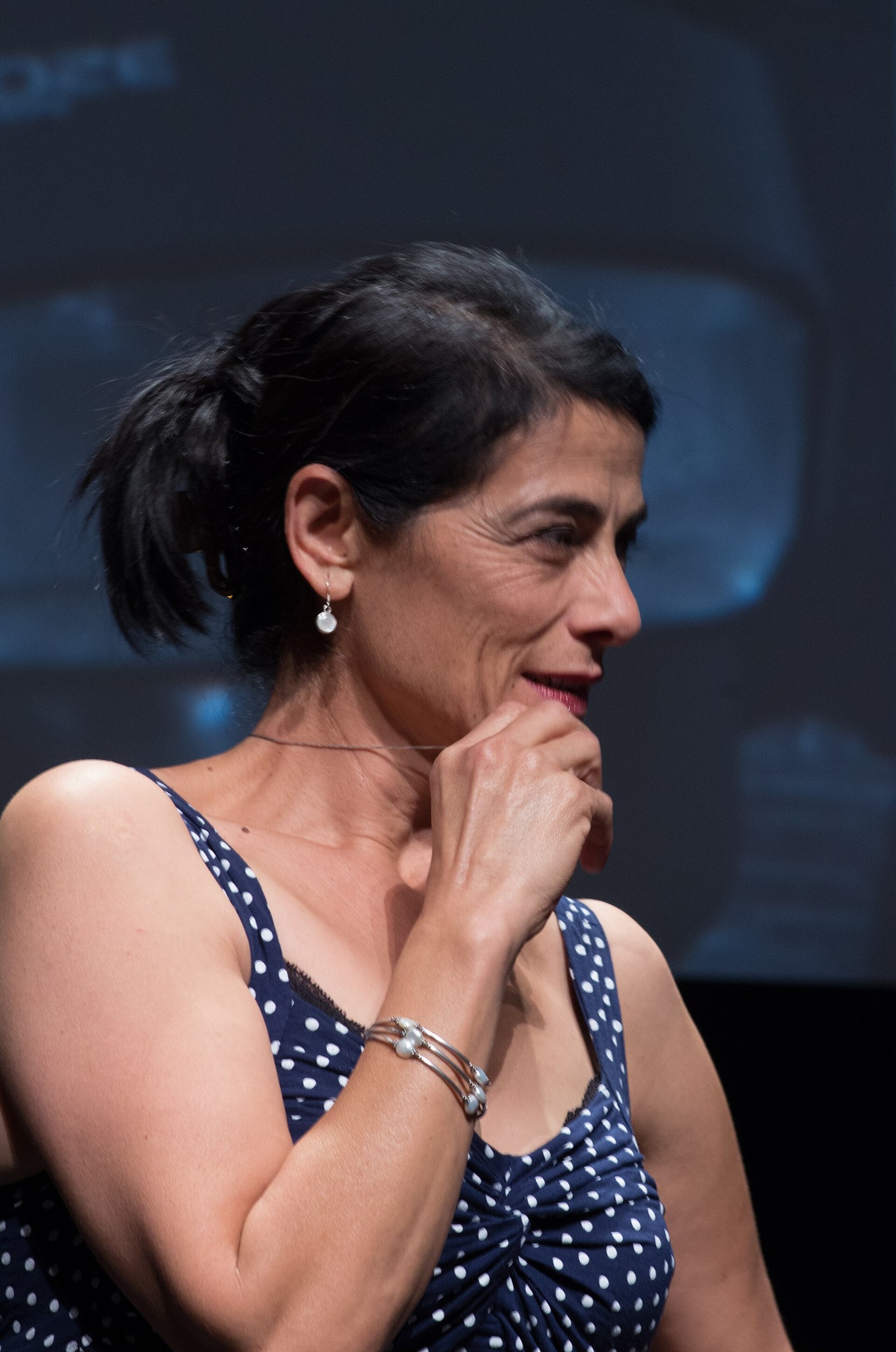 Woman with dark hair in a ponytail and a navy polka-dot top smiles, hand at her chin, on a stage with a dark blue backdrop