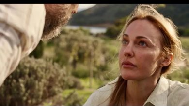 Film still of a woman with blond hair looking up at a bearded man in the foreground, with olive trees and hills behind.