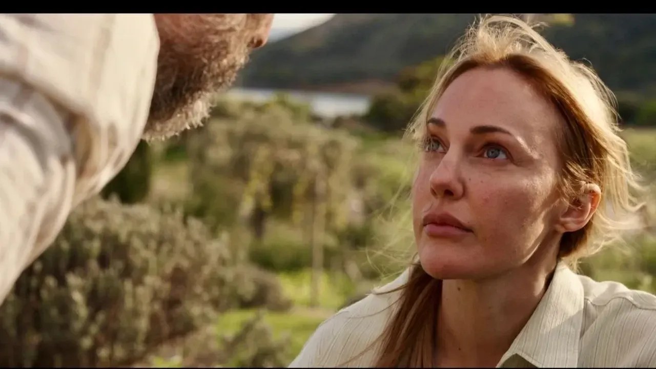 Film still of a woman with blond hair looking up at a bearded man in the foreground, with olive trees and hills behind.