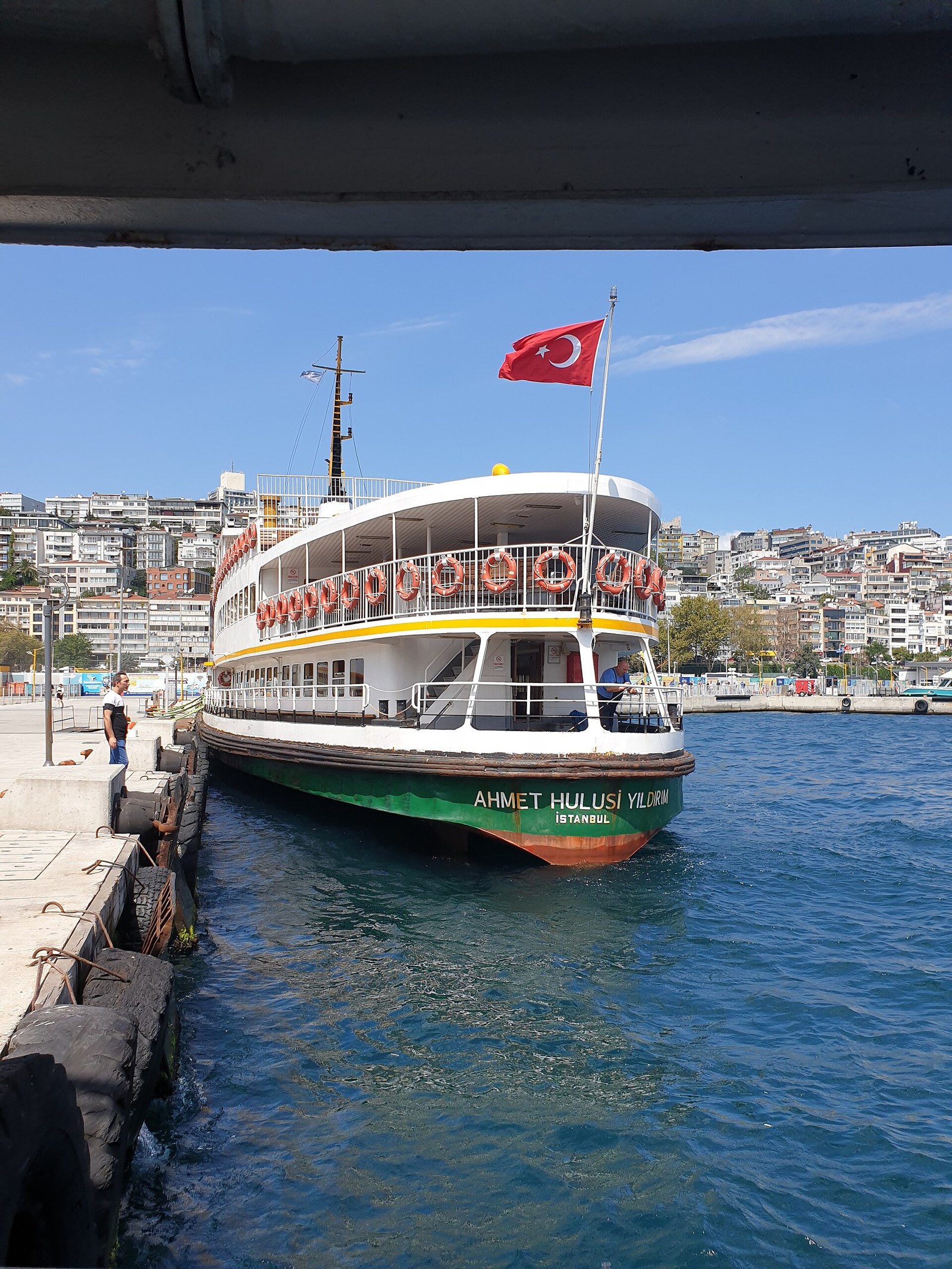 Istanbul ferry Ahmet Hulusi Yildirim moored at a Bosphorus pier, a Turkish flag flying and hillside buildings behind