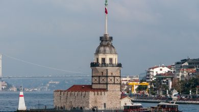 Maiden's Tower on its islet in the Bosphorus, Istanbul, with a Turkish flag, two small boats and a suspension bridge behind.