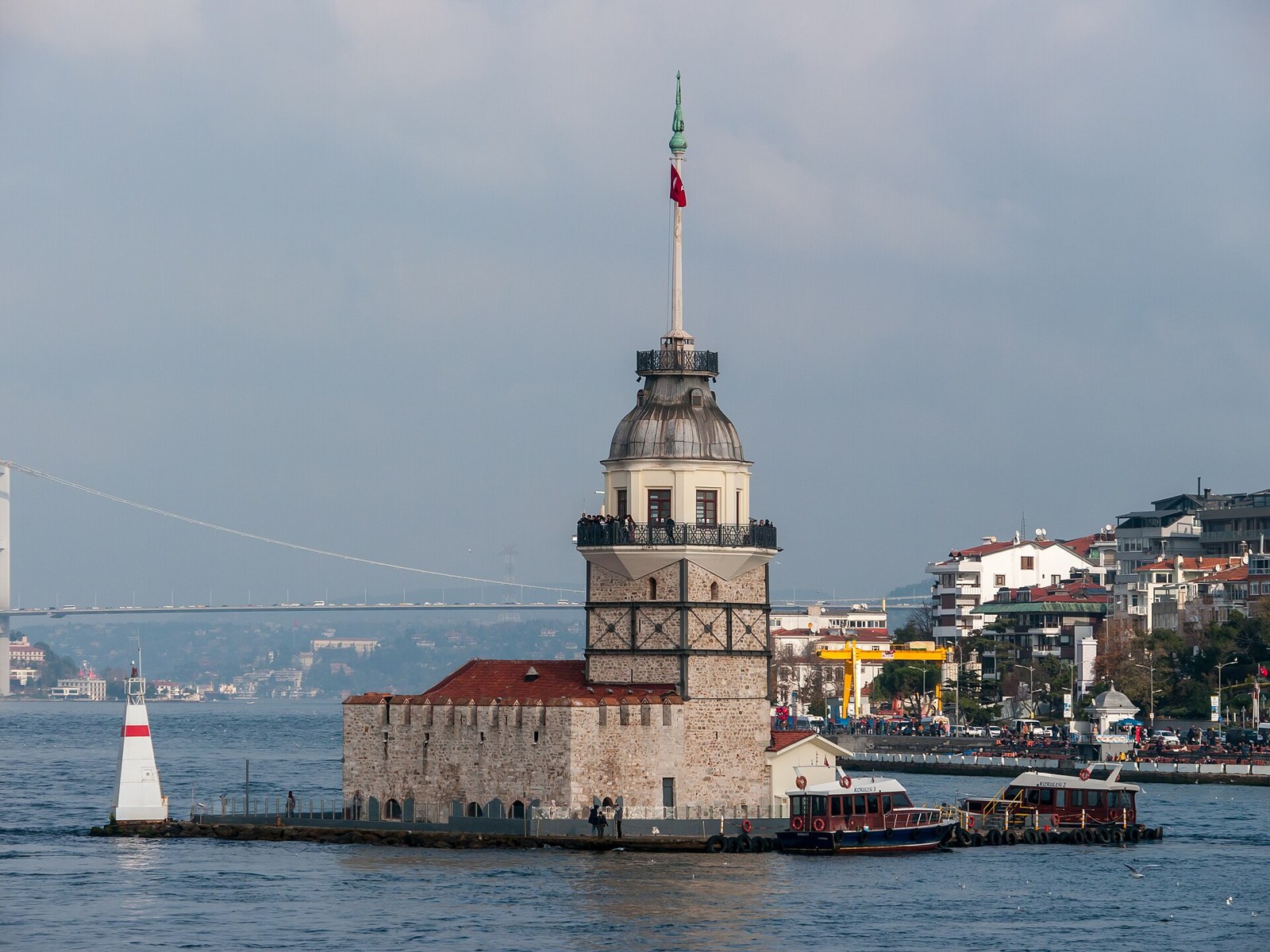 Maiden's Tower on its islet in the Bosphorus, Istanbul, with a Turkish flag, two small boats and a suspension bridge behind.