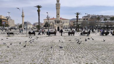 Izmir Clock Tower in Konak Square, with pigeons on the cobblestones, people on benches, palm trees and a mosque behind.