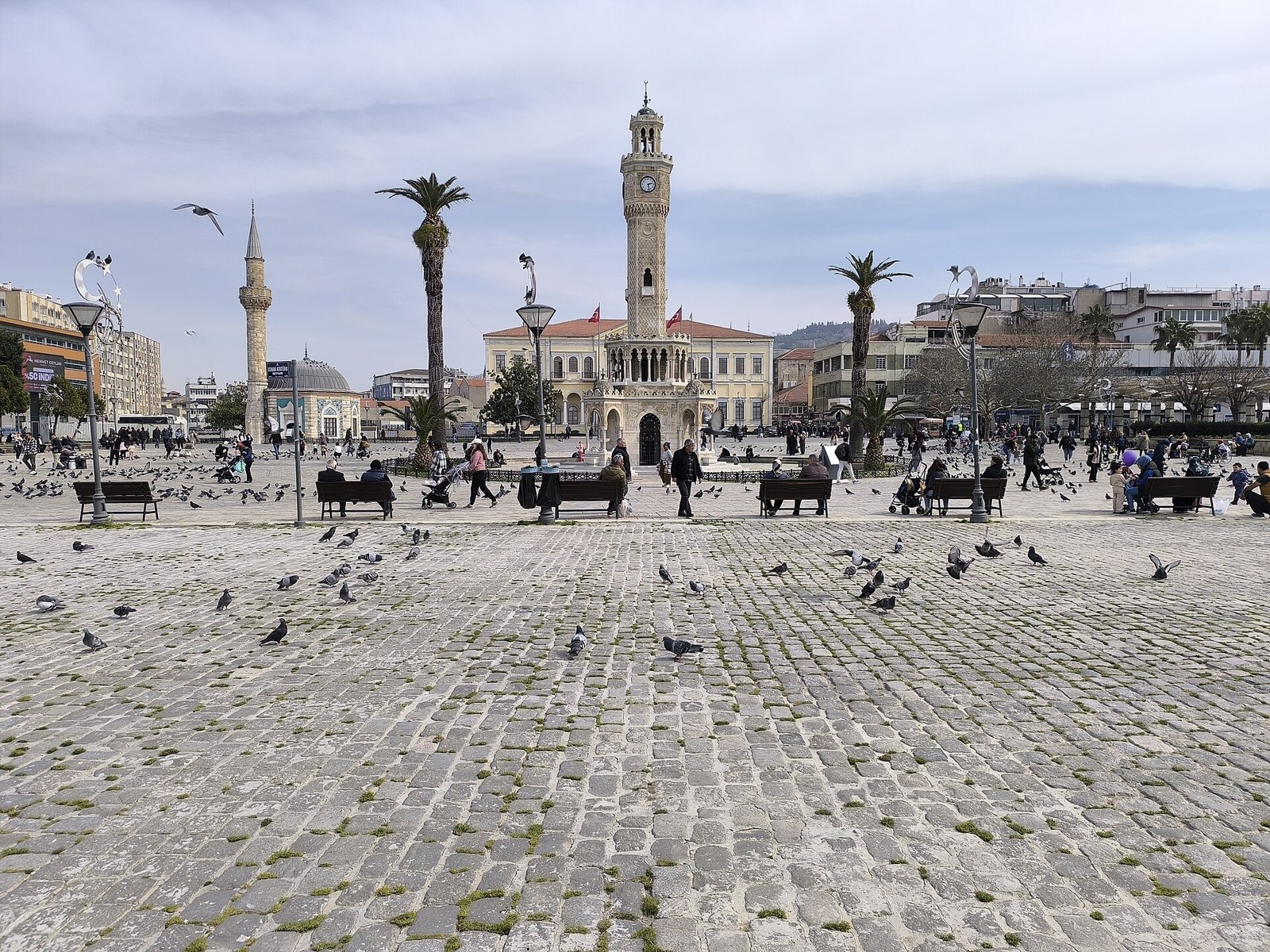 Izmir Clock Tower in Konak Square, with pigeons on the cobblestones, people on benches, palm trees and a mosque behind.