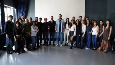 Large group of actors and crew members posing together in a bright studio room with a blank white screen behind them