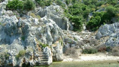 Rock-cut stone steps on a rocky, scrub-covered Mediterranean shore beside clear turquoise water, near Kekova, Turkey