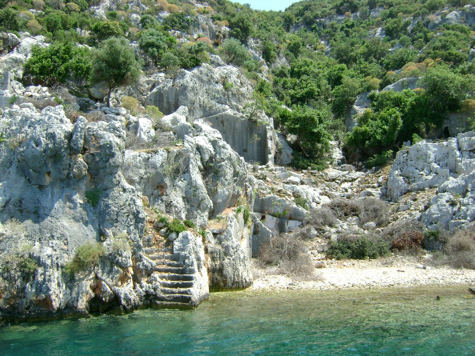 Rock-cut stone steps on a rocky, scrub-covered Mediterranean shore beside clear turquoise water, near Kekova, Turkey
