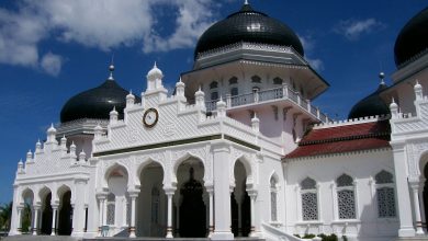 White mosque with black domes, ornate arches and a clock facade under a blue sky, resembling Baiturrahman Grand Mosque in Aceh