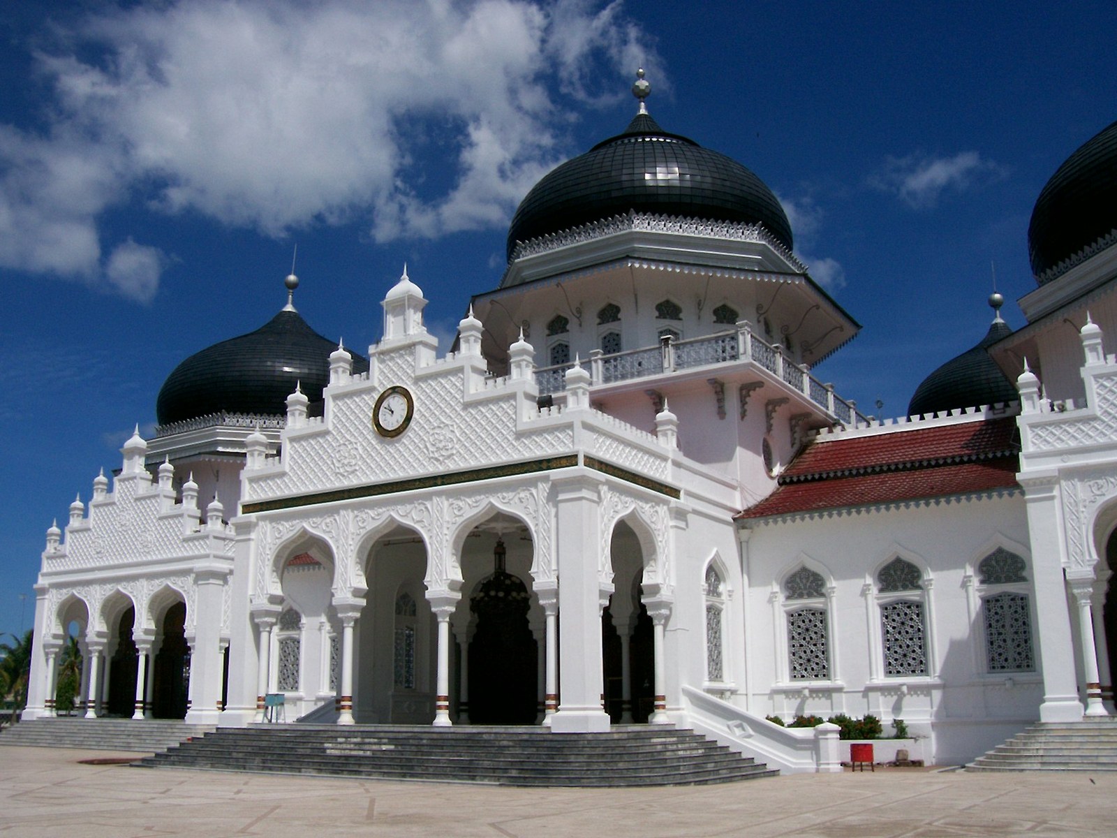 White mosque with black domes, ornate arches and a clock facade under a blue sky, resembling Baiturrahman Grand Mosque in Aceh