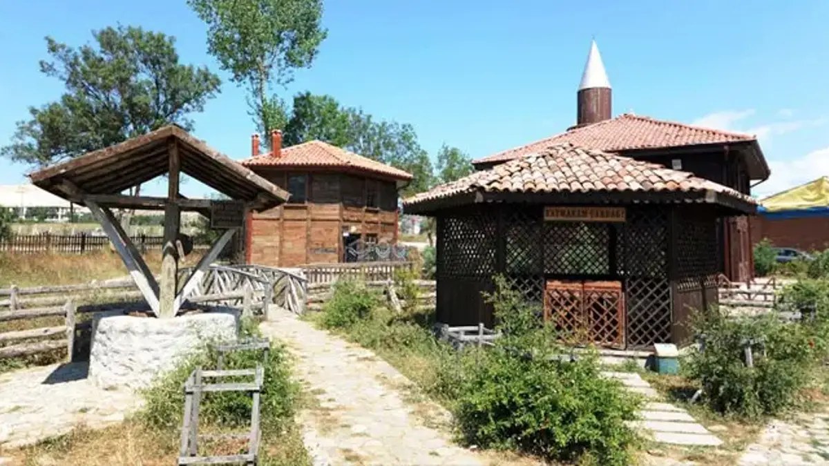 Wooden mosque with a tiled roof and white-capped minaret beside a wooden well canopy and a stone path on a sunny day
