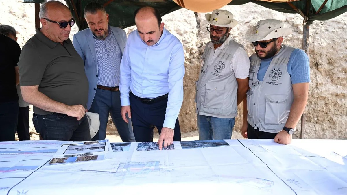 Officials and archaeologists in vests and hats study site plans on a table at an excavation site beside an earthen wall.