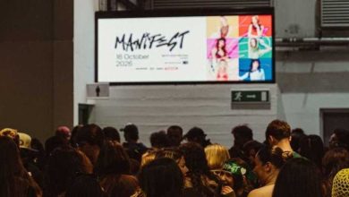 Crowd of fans queue beneath a screen showing Manifest, 16 October 2026, with six colourful portraits of the group members.